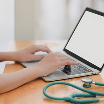 doctor typing on a laptop with a stethoscope curled about beside the keyboard, and a patient skyping or zooming for a telehealth consult 